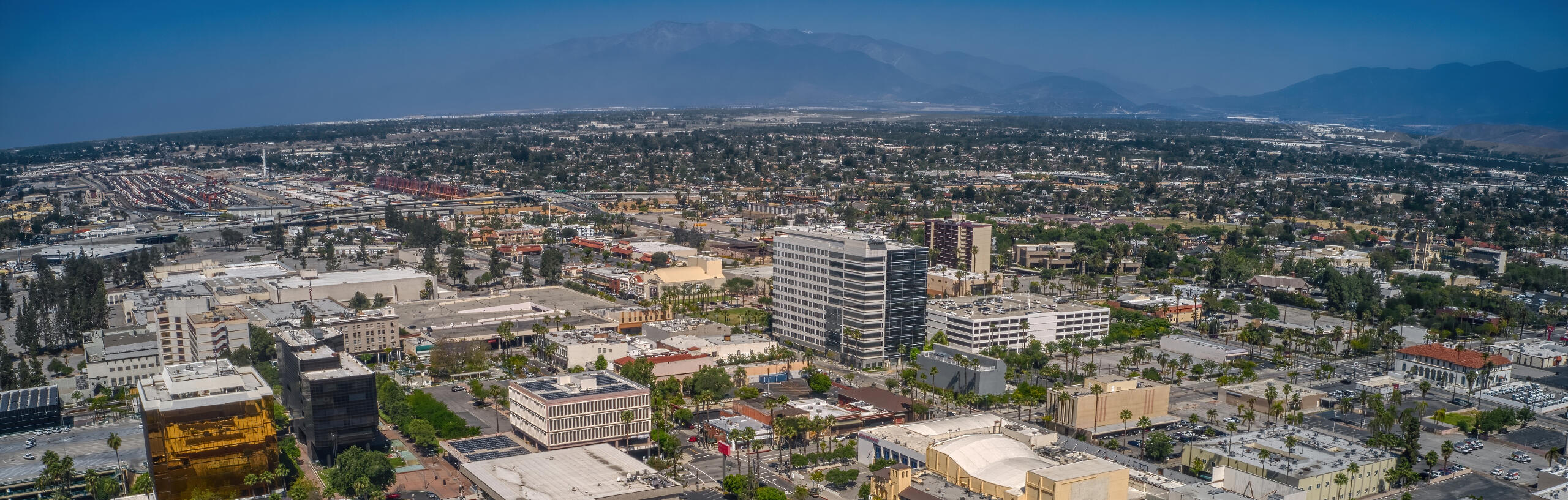 Aerial view of a cityscape with a mix of low-rise and high-rise buildings. The scene includes streets, trees, and parking lots, with distant mountains in the background and clear blue skies overhead.